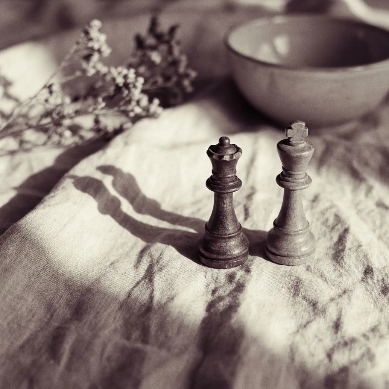 Two chess pieces, a queen and a king, standing close together on cream linen in soft window light.