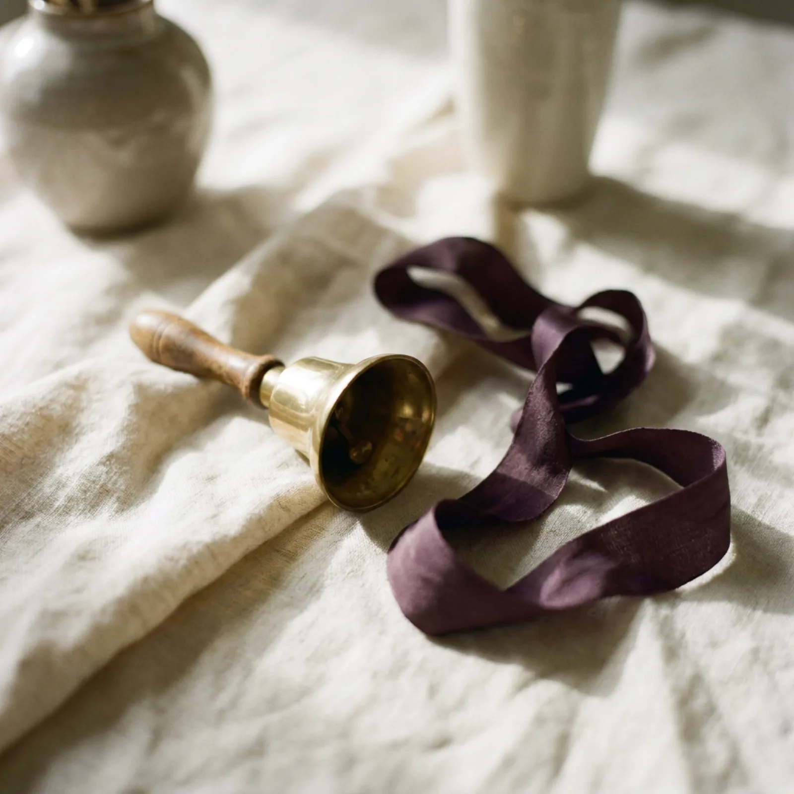 A small brass handbell beside a knotted plum silk ribbon on cream linen in soft window light, natural still-life arrangement.